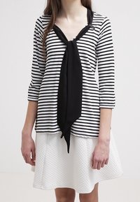 Randig svart och vit topp med en halsknut, i kombination med en texturerad vit kjol. Har trekvartsärmar och en avslappnad passform.