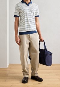 Pruhované polo tričko v krémové a námořnické modři s krátkým rukávem, spárované s světlými béžovými kalhotami. Drží námořnickou tašku a má na sobě černé boty s hrubou podrážkou.