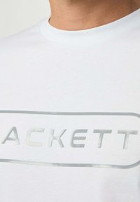 Camiseta de algodón blanca con un escote redondo y un logo plateado y reflectante que dice "ACKETT" en un contorno rectangular a lo largo del pecho.