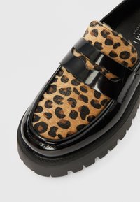 Mokasína s leopardím vzorom s lesklými čiernymi detailmi z lakovanej kože, vybavená ryhovanou čiernou podrážkou a mäkkým textúrovaným zvrškom pripomínajúcim srsť.