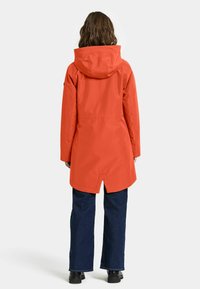 Vandafvisende orange regnjakke med hætte, præget af en snævret talje og en let længere ryg, parret med mørkeblå lige ben jeans.