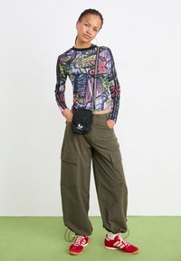 Långärmad, flerfärgad grafisk tröja med texten "Adidas", i kombination med olivgröna cargobyxor och röda sportskor. Liten svart crossbody-väska ingår.