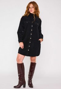 Abito camicia in velluto nero con maniche lunghe, chiusura con bottoni, due tasche sul petto e tasche laterali. Abbinato a stivali bordeaux fino al ginocchio.