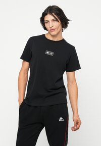 Camiseta de algodón negra con cuello redondo, mangas cortas y un pequeño parche de logo blanco en la parte superior del pecho. Corte casual.