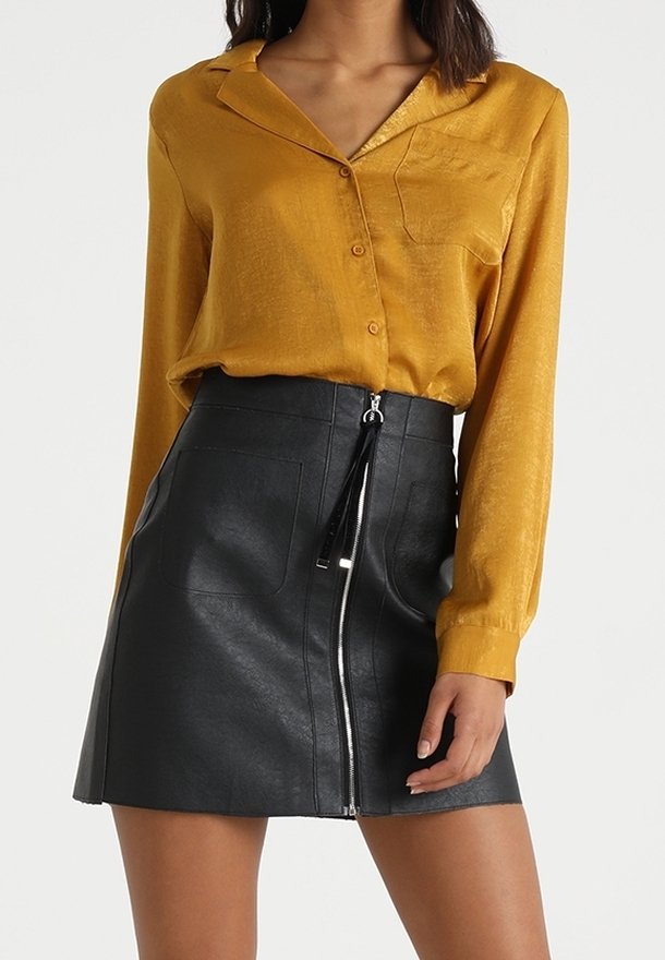Blouse en soie moutarde à manches longues et poche, associée à une mini-jupe en cuir noir avec une fermeture éclair frontale et deux poches.