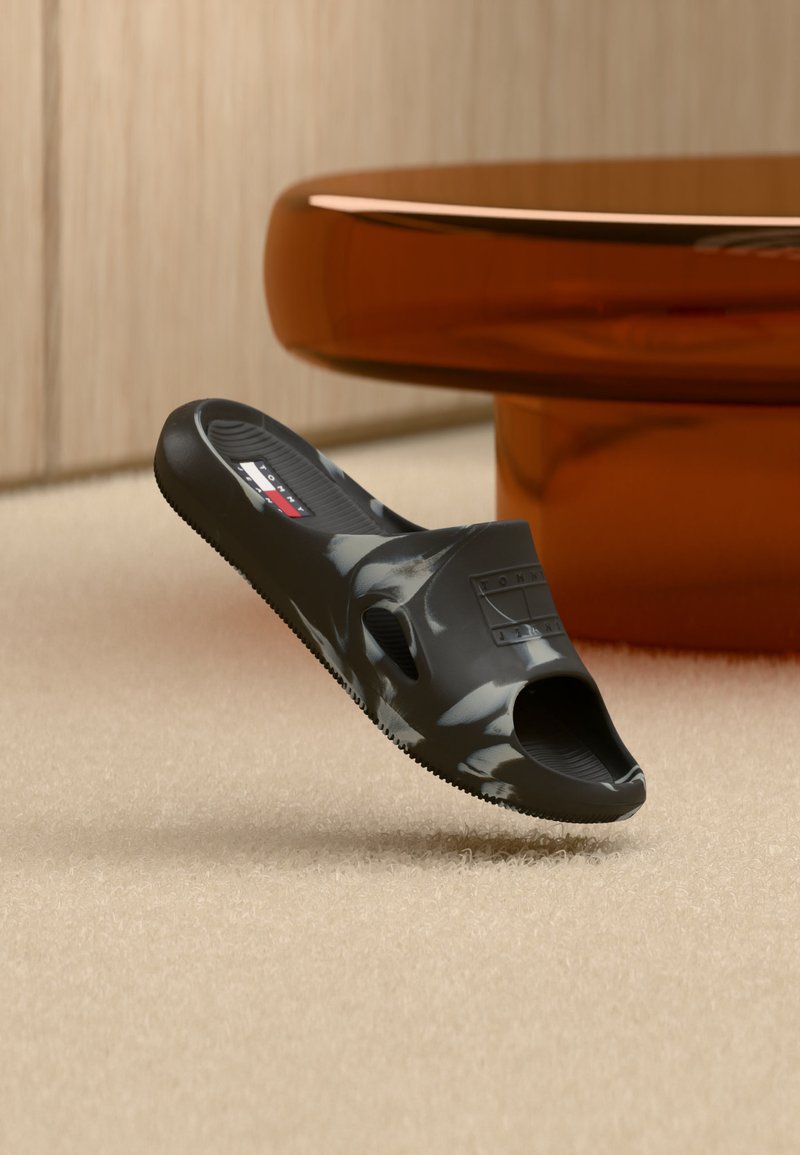 Μαύρο και λευκό μαρμάρινο σαγιονάρι με λογότυπο Tommy Hilfiger σε μπεζ χαλί, κοντά σε στρογγυλό τραπέζι κεχριμπαρένιου χρώματος.