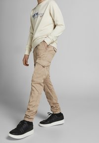 Person i beige cargobyxor med fickor, krämfärgad sweatshirt med logotyp, svarta sneakers med vita sulor, går mot en grå bakgrund.