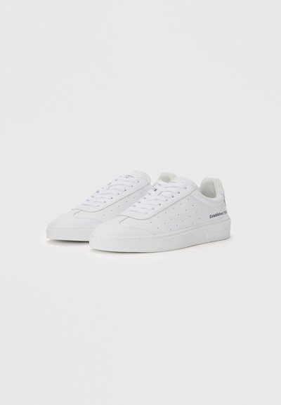 Baskets blanches avec un design perforé, lacets plats, matière douce et semelle texturée ; logo sur le talon et le côté.