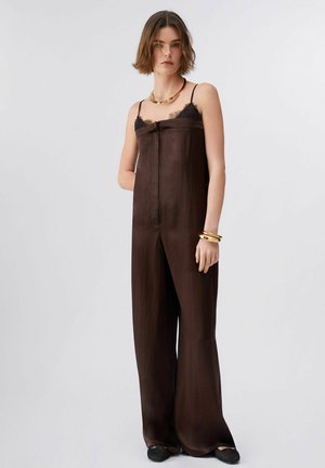 Frau trägt einen ärmellosen braunen Overall mit Spitzenbesatz, goldener Halskette und Armbändern, steht vor einem schlichten hellen Hintergrund.