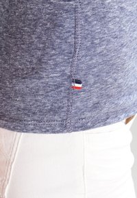 Blå melerad bomullströja med en detalj i sömmen. Har en liten röd, vit och blå logotagg vid fållen. Styler med vita byxor.