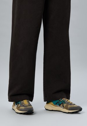 Legs wearing black wide-leg pants and multicolored sneakers with beige, green, yellow, and black accents on a light gray background.