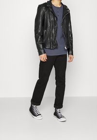 Mężczyzna w czarnej skórzanej kurtce na zamek, ciemnoniebieskiej koszulce henley, czarnych spodniach z mankietami i czarnych sneakersach za kostkę z białymi sznurówkami.