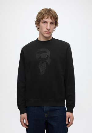 Młody mężczyzna o jasnobrązowych falowanych włosach nosi czarną bluzę z subtelną, wypukłą postacią z kreskówki w okularach przeciwsłonecznych oraz niebieskie dżinsy.