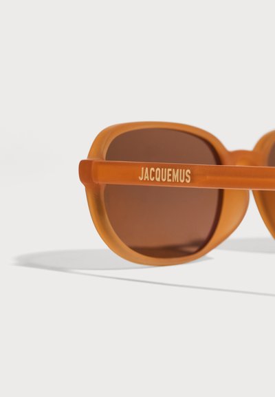 Оранжеви полупрозрачни слънчеви очила с кафяви лещи и "JACQUEMUS" написано на страничната рамка с бели букви.