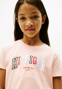 Lyserød bomulds T-shirt med fed hvid, rød og marineblå "HILFIGER" tekst; yderligere "NEW YORK CITY U.S.A." detalje nedenfor. Korte ærmer.