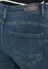 Jeans de mezclilla azul oscuro con un diseño clásico de cinco bolsillos, costuras visibles y una etiqueta de cuero marrón con "ONLY" en la cintura.