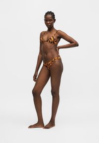 Γυναίκα που στέκεται ξυπόλητη φορώντας ένα πορτοκαλί και μαύρο μπικίνι με σχέδιο ζώου, με το ένα χέρι στη μέση, πάνω σε λευκό φόντο.