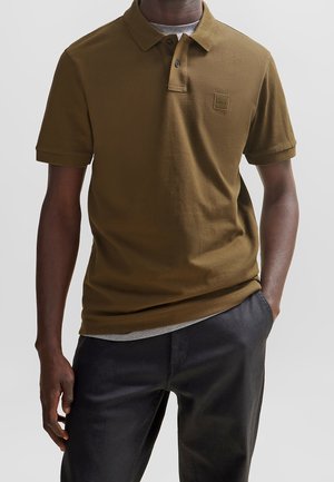 Olivgrünes Poloshirt mit klassischem Kragen und kurzen Ärmeln, versehen mit einem kleinen Logopatch auf der Brust, kombiniert mit schwarzen Hosen.
