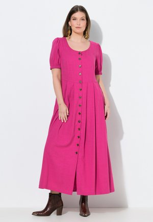 Vrouw die een felroze midi-jurk met knopen aan de voorkant en pofmouwen draagt, gecombineerd met bruine enkellaarsjes, staand tegen een witte achtergrond.