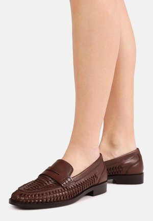 Nogi w ciemnobrązowych plecionych skórzanych mokasynach z zamkniętymi piętami i niskimi czarnymi podeszwami na jasnoszarym tle.