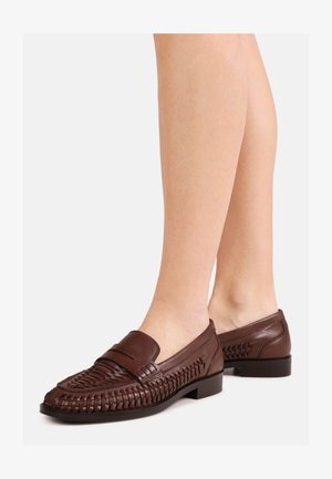 Benen met donkerbruine geweven leren loafers met gesloten hielen en lage zwarte zolen op een lichtgrijze achtergrond.