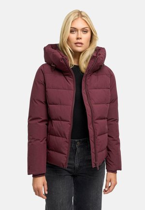 Donna bionda che indossa una giacca imbottita bordeaux con cappuccio, top nero e jeans scuri, in piedi davanti a uno sfondo chiaro e uniforme.