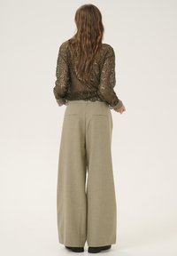 Top de encaje oliva transparente con mangas largas, combinado con pantalones beige de pierna ancha. Patrón texturizado y detalle de cintura ajustada en la parte superior.