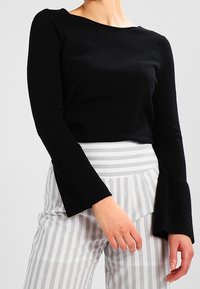 Musta pitkähihainen paita leveällä venepääntiellä ja kellohihansuilla, yhdistetty harmaa-valkoisiin raidoitettuihin korkeavyötäröisiin housuihin.