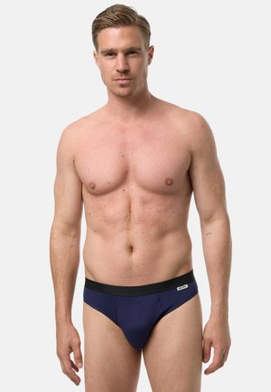 Mężczyzna bez koszulki o krótkich blond włosach, ubrany w granatowe slipy z czarnym paskiem, stojący na tle jasnego, jednolitego tła.