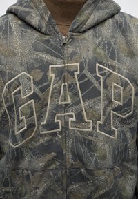 Sweat à capuche zippé camouflage avec de grandes lettres en contour formant GAP sur le devant, porté sur un t-shirt marron à col rond.