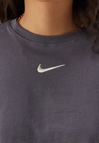 Tmavě šedé bavlněné tričko s kulatým výstřihem a malým bíle vyšitým logem Nike Swoosh na hrudi. Jemná textura.