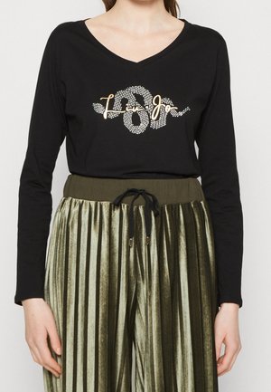 Frau trägt ein schwarzes langärmeliges V-Ausschnitt-Oberteil mit goldenem und strassbesetztem "Liu Jo"-Logo sowie olivgrüne plissierte Samthose mit Kordelzugbund.