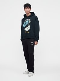 Άνδρας που φοράει μαύρο φούτερ Moschino με γραφικό μιας χήνας που κρατάει ομπρέλα, μαύρη φόρμα Moschino και λευκά αθλητικά παπούτσια, στέκεται μπροστά σε λευκό φόντο.