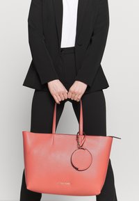 Sac fourre-tout rose corail avec finition en cuir texturé, double anses, et un pendentif circulaire attaché. Présente un marquage subtil sur le devant.
