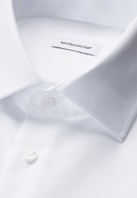 Weißes Hemd mit weiter Kragenform, sichtbarem oberstem Knopf geschlossen und "seidensticker" Markenetikett innen am Kragen.