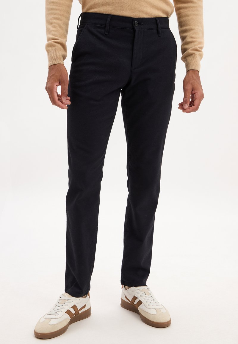Pantalons slim-fit bleu marine en coton avec une texture lisse, dotés de poches avant et d'une fermeture à bouton, associés à des baskets blanches.