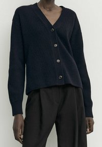 Gilet côtelé bleu marine avec un col en V, comportant six boutons à l'avant et des manches longues. Porté par-dessus un pantalon sombre, mettant en avant la texture et l'ajustement.