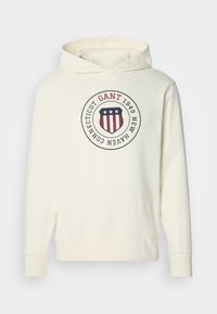 Sweatshirt cu glugă de culoare albă-crem, cu un logo circular în formă de scut, care include stele și dungi, și textul "GANT 1949 New Haven Connecticut" pe piept.