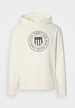 Sweat à capuche blanc cassé avec un logo en forme de bouclier circulaire arborant des étoiles et des rayures, ainsi que le texte "GANT 1949 New Haven Connecticut" sur la poitrine.