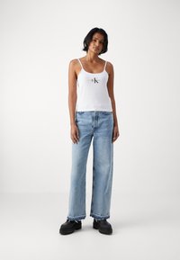 Hvit tanktopp med tynne stropper som har en svart logo. Kombinert med lyseblå vid jeans og svarte sko med tykk såle.
