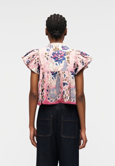 Personne avec un chignon tressé porte un chemisier à manches courtes à motif floral avec des épaules à volants et un pantalon foncé avec des poches arrière, debout de dos.