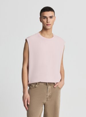Giovane uomo con capelli corti e scuri che indossa una canotta rosa pallido e pantaloni beige, in piedi davanti a uno sfondo grigio chiaro.