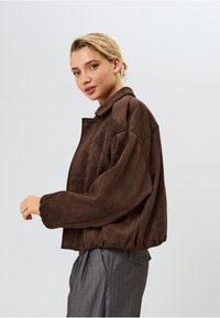 Rudas zomšinės trumpa striukė su apykakle, plačiomis rankovėmis, elastinga apatine kraštine ir subtiliu prekės ženklu krūtinėje. Tekstūruota ir minkšta išvaizda.