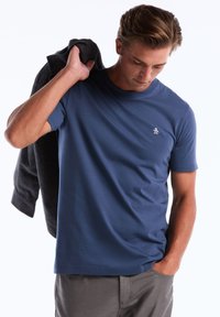 T-shirt à manches courtes bleu marine en tissu lisse, avec un col rond et un logo de pingouin sur la poitrine gauche. Une veste grise est drapée sur l'épaule.