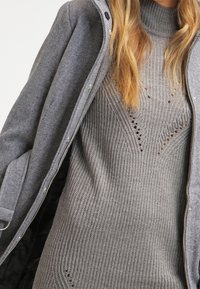 Šedé žebrované pletené šaty se složitými vzory a výřezy, vrstvené pod šedým kabátem na zip s texturovanou tkaninou.