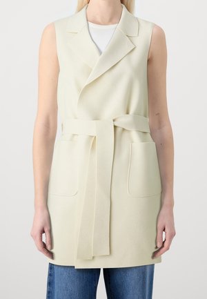 Sleeveless light beige belted vest with lapel collar and two square front pockets worn over a white top and blue jeans.