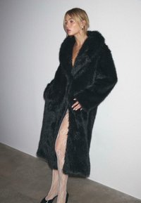 Crni kaput od umjetnog krzna s velikim ovratnikom, otvorenim prednjim dijelom, dva bočna džepa i dugim rukavima. Model nosi čipkast bodi ispod.