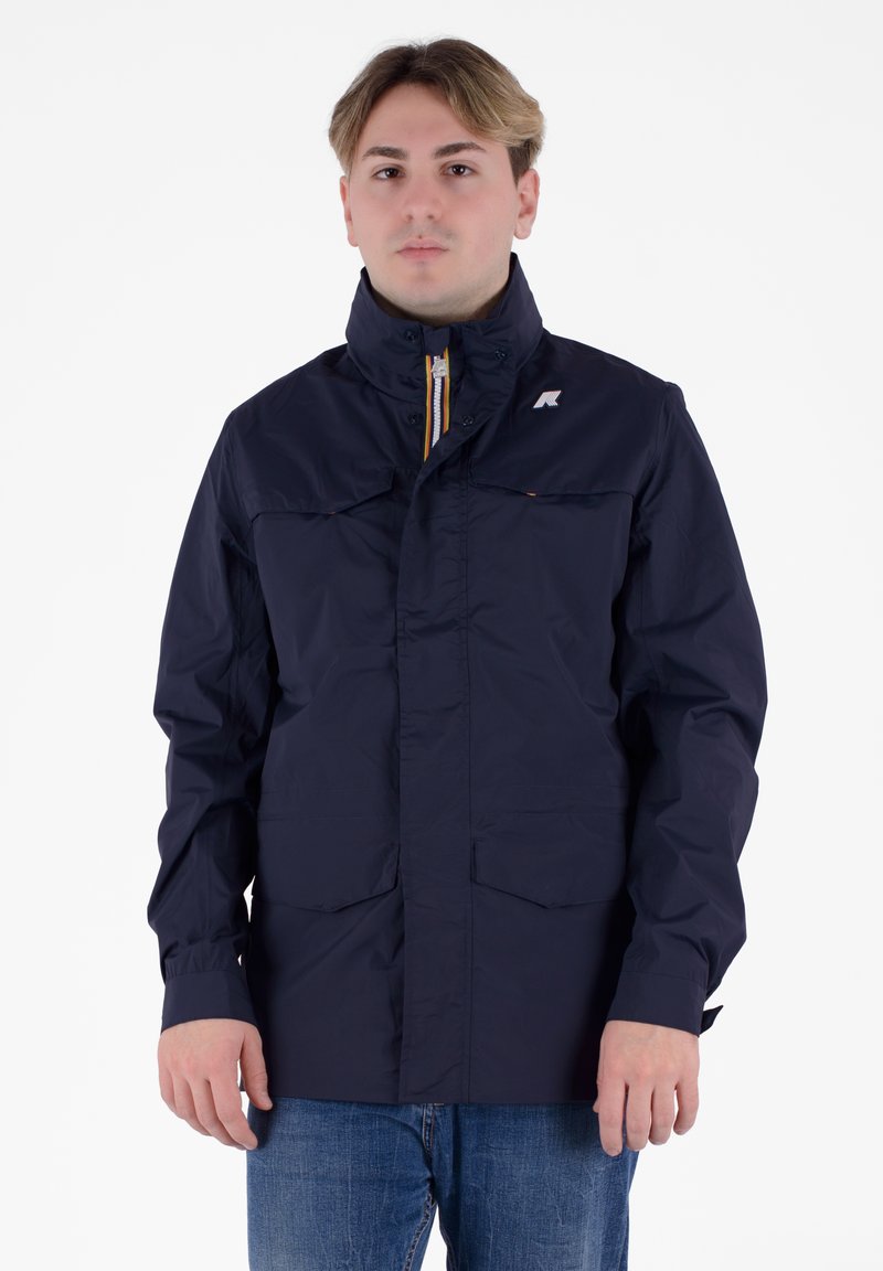 Giacca impermeabile blu navy con colletto alto, zip frontale, due tasche sul petto e accento del logo. Tessuto liscio e vestibilità comoda.