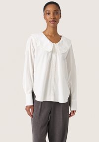 Blusa bianca a maniche lunghe con un grande colletto arricciato; tessuto leggero; chiusura a bottoni sul davanti; indossata con pantaloni grigi a righe.