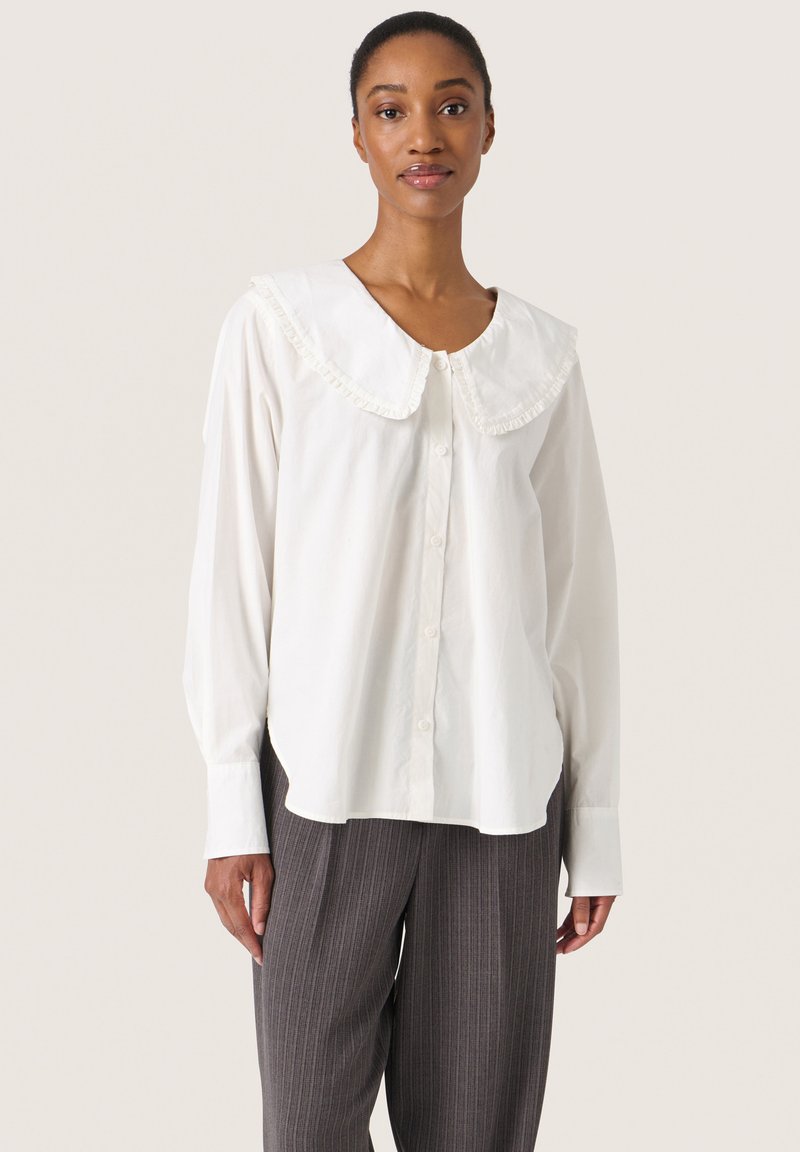 Blusa bianca a maniche lunghe con un grande colletto arricciato; tessuto leggero; chiusura a bottoni sul davanti; indossata con pantaloni grigi a righe.